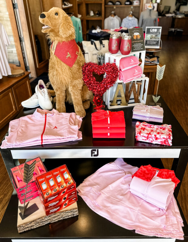 Valentine’s-themed golf shop display featuring coordinated pink and red apparel folded on a black merchandise table, accented with heart décor, a plush golden retriever prop wearing a red bandana, insulated tumblers, golf balls, and gift items arranged for a holiday promotion.