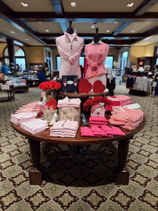 Round display table inside a golf shop featuring coordinated pink apparel, folded sweaters, mannequins in pastel tops, red floral accents, and gift items arranged for a themed retail presentation.