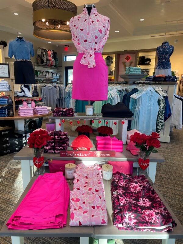 Valentine’s-themed golf shop display featuring a mannequin in a pink heart-print top and bright pink skirt, surrounded by coordinated folded apparel, red rose accents, heart string lights, and a “Valentine Forever” sign centered on tiered merchandise tables.