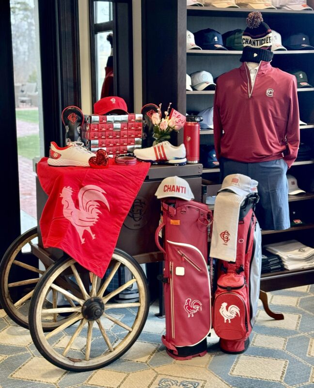 A "Chanticleer" themed retail display at Greenville Country Club features a wooden cart with large spoked wheels. A mannequin wears a maroon quarter-zip and a "CHANTICLEER" pom-pom beanie. The setup includes red and white golf bags with rooster logos, stacks of red TaylorMade ball boxes, and white Adidas golf shoes.