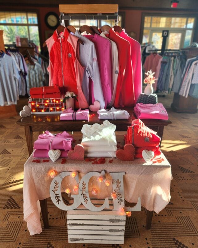 Valentine’s-themed golf shop display featuring coordinated pink, red, and white apparel on a wooden table, accented with heart décor, soft lighting, folded sweaters, and a decorative “GCL” sign at the base.