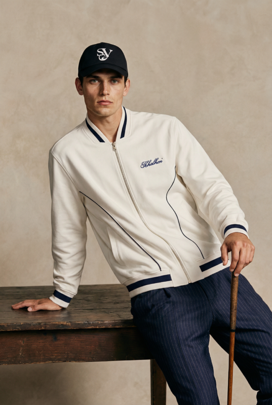 Male model wearing a cream zip-up golf jacket with navy trim and Khalhon logo, paired with navy pinstripe pants and a black cap, leaning against a wooden table while holding a golf club.