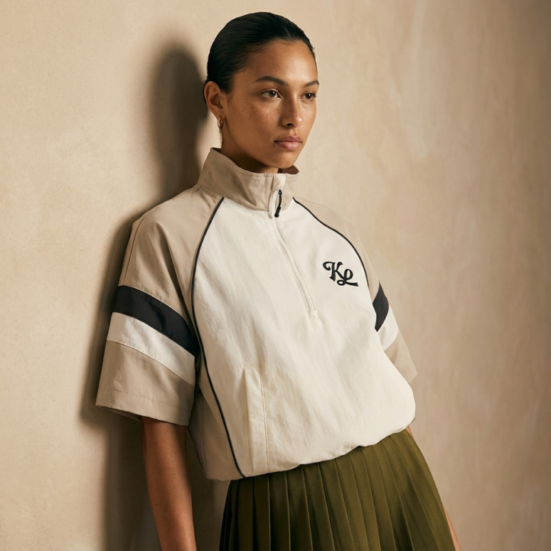 Female model wearing a cream short-sleeve quarter-zip golf top with black accent stripes and logo, paired with an olive pleated skirt, leaning against a textured studio wall.