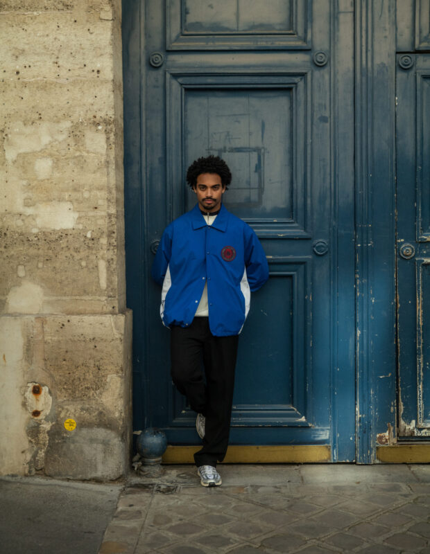 Male model wearing a bright blue golf-inspired jacket with logo detailing, black pants, and sneakers, standing with hands behind his back against a weathered blue door and stone exterior in an urban setting.