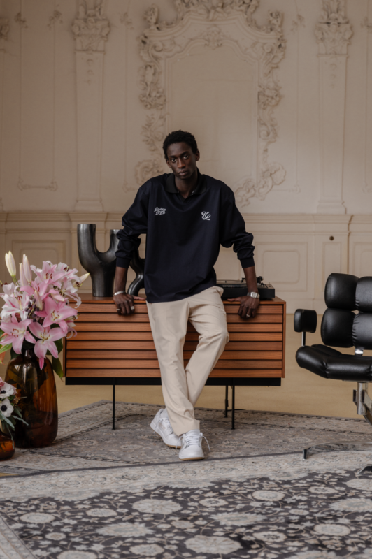 Male model wearing a navy long-sleeve golf pullover with embroidered logo and light khaki pants, leaning against a wooden console table in an elegant, vintage-style interior with floral arrangements and modern seating.