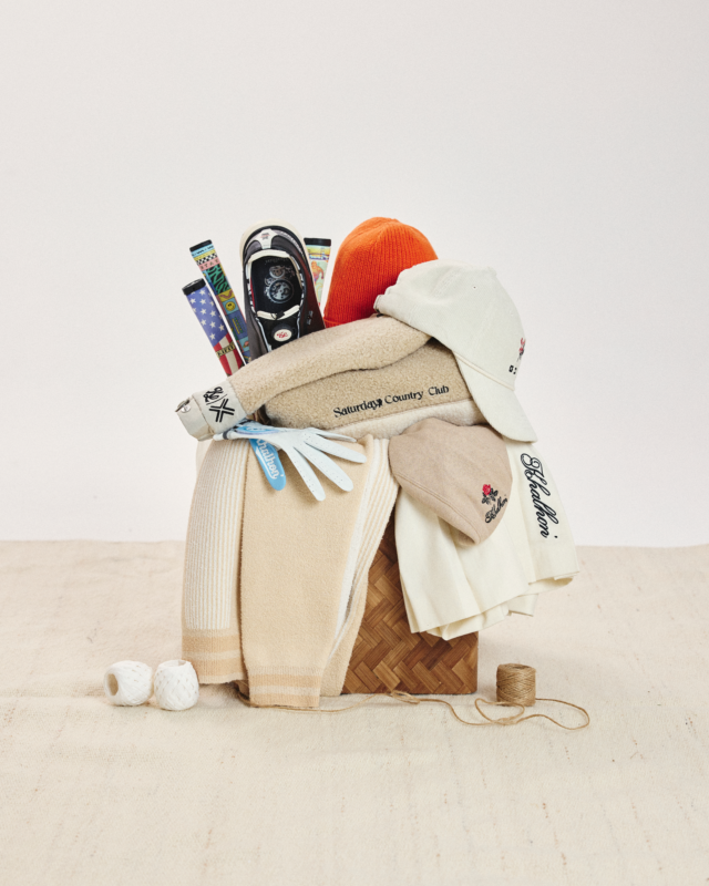 Curated still life of Khalhon golf accessories and apparel, including headcovers, gloves, knit hats, towels, and folded garments, styled atop a woven box on a neutral studio backdrop.