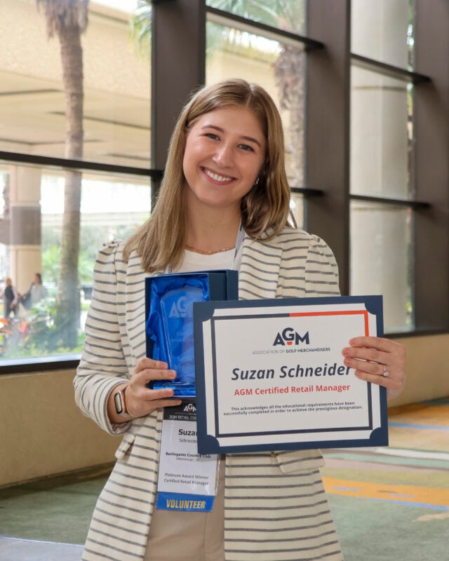 A smiling woman, Suzan Schneider, holding an AGM Certified Retail Manager certificate and a glass award. She is wearing a white and grey striped blazer and a name tag.