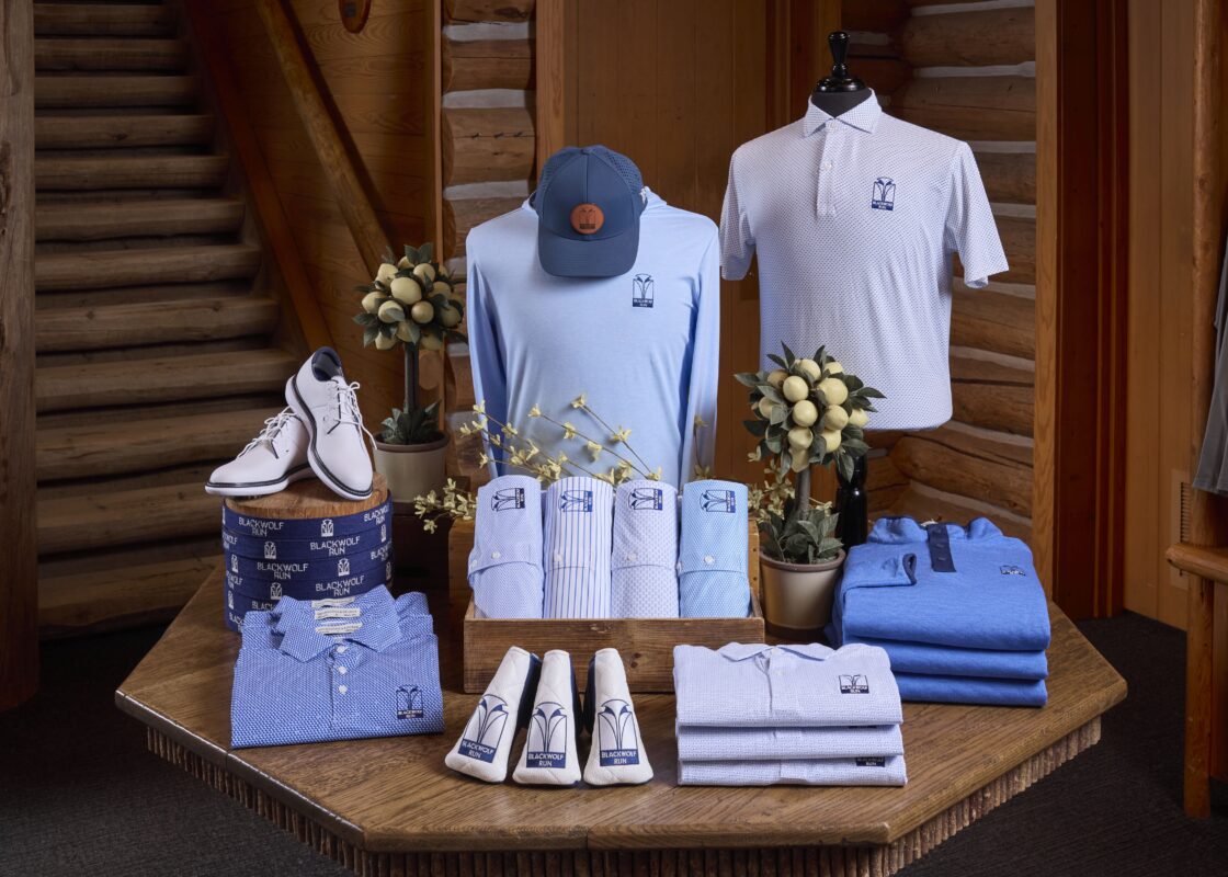 A curated men’s apparel vignette at Blackwolf Run featuring a blue color story with logo-branded performance polos, hoodies, headwear, and coordinated accessories on a rustic wooden display table.