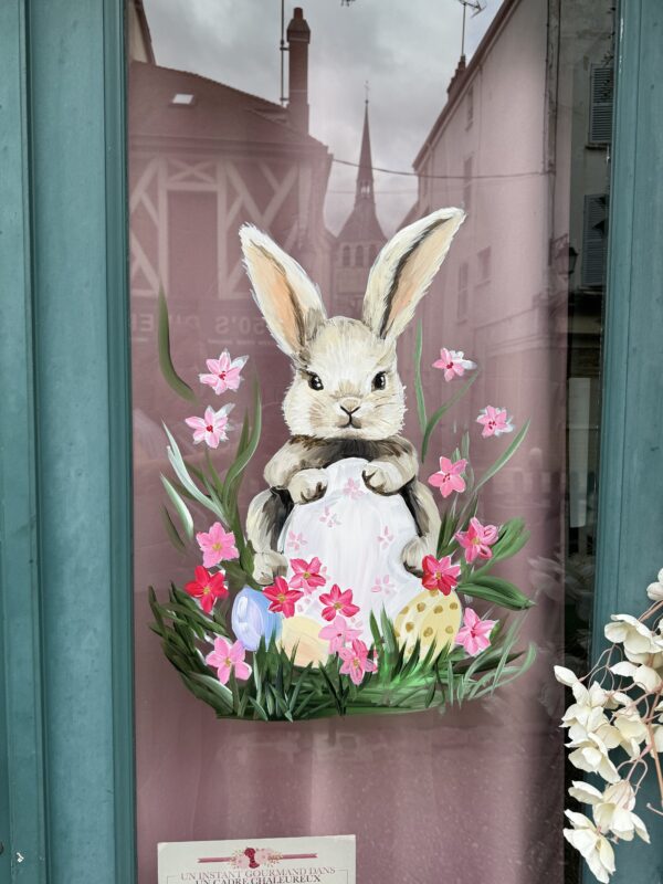 A hand-painted illustration on a glass window depicts a fluffy brown and white Easter bunny holding a large white egg.