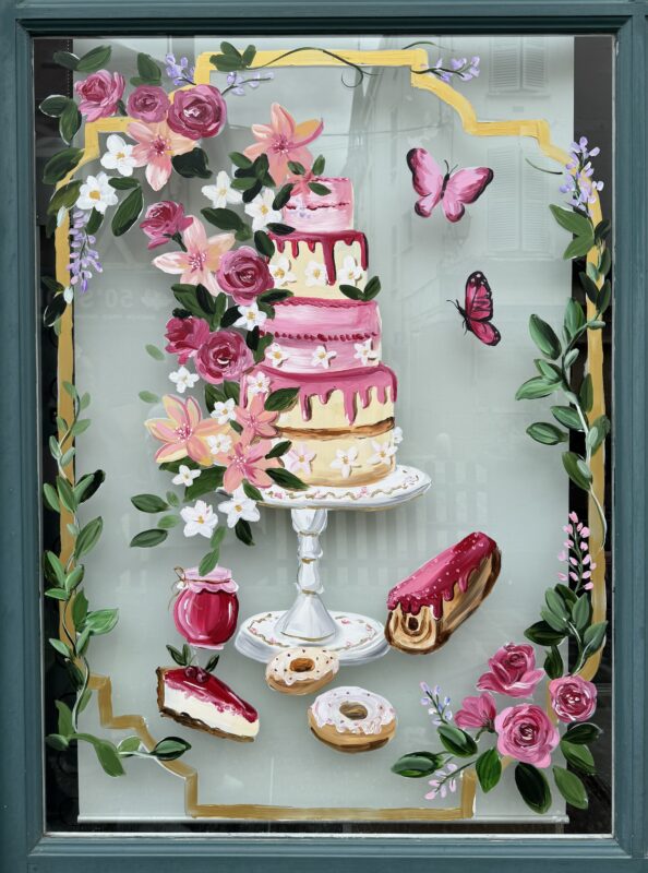 Storefront window painting of a 4-tiered cake, various other sweet treats, and flowers.