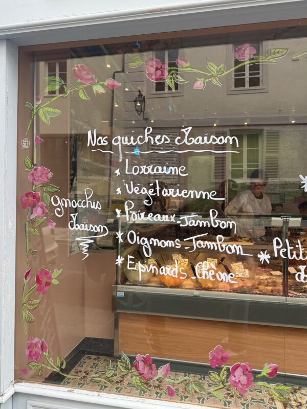 Window of a French bakery with the words (translated from French) "Our Seasonal Quiches: Lorraine, Vegetarian, Leeks & Ham, Onions & Ham, Spinach & Goat Cheese. Seasonal Gnocchi" handwritten on the window. There are painted flowers around the menu.