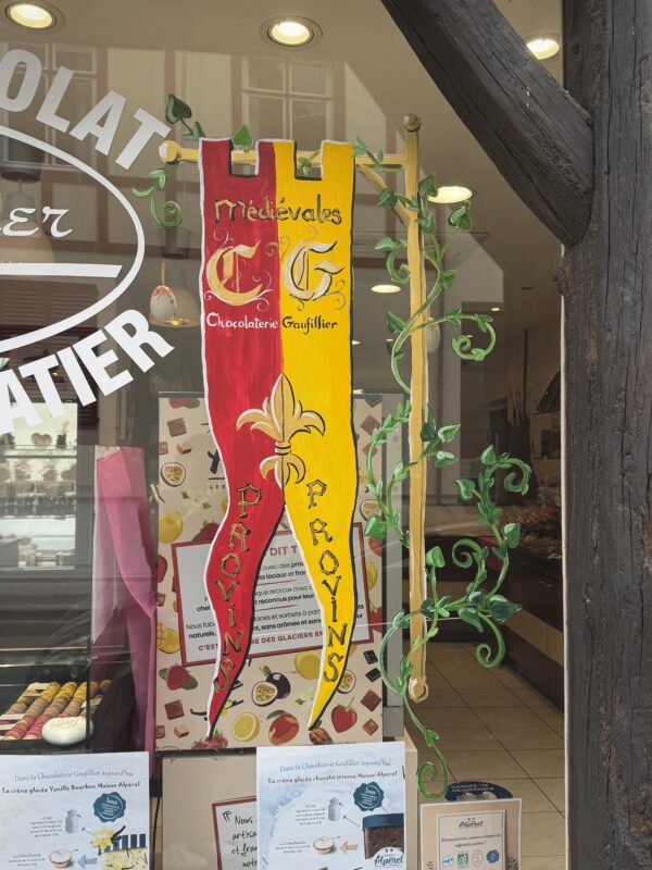 A hand-painted vertical banner on a glass window, split down the middle in red and yellow. The red side features a golden "C" and the yellow side a golden "G," representing "Chocolaterie Gaufillier." Below the initials is a gold fleur-de-lis and the word "PROVINS" written vertically on both halves.