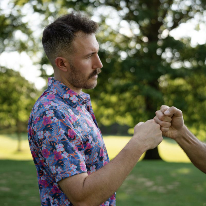 Golfer wearing a colorful floral polo fist-bumping another player on a golf course, with trees and soft sunlight in the background.