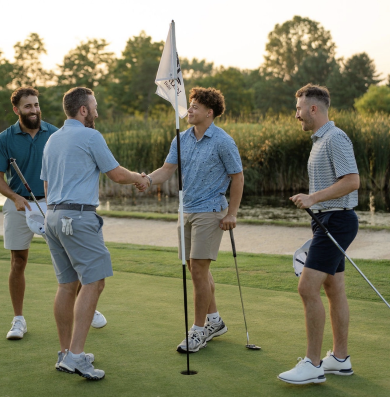 Four golfers standing on a putting green at sunset, shaking hands beside the flagstick while holding their putters, with tall grass and water in the background.