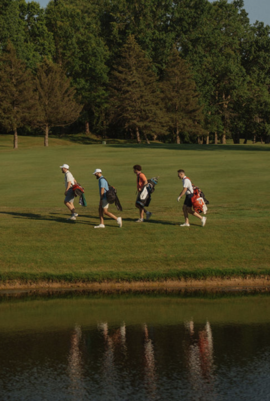 Four golfers walking in a line across a green fairway carrying their golf bags over their shoulders, with trees and late afternoon light in the background.