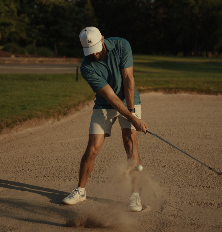 Golfer wearing a teal polo, light shorts, and a white cap with a Highland cow logo hitting a bunker shot, with sand spraying as the club makes contact.