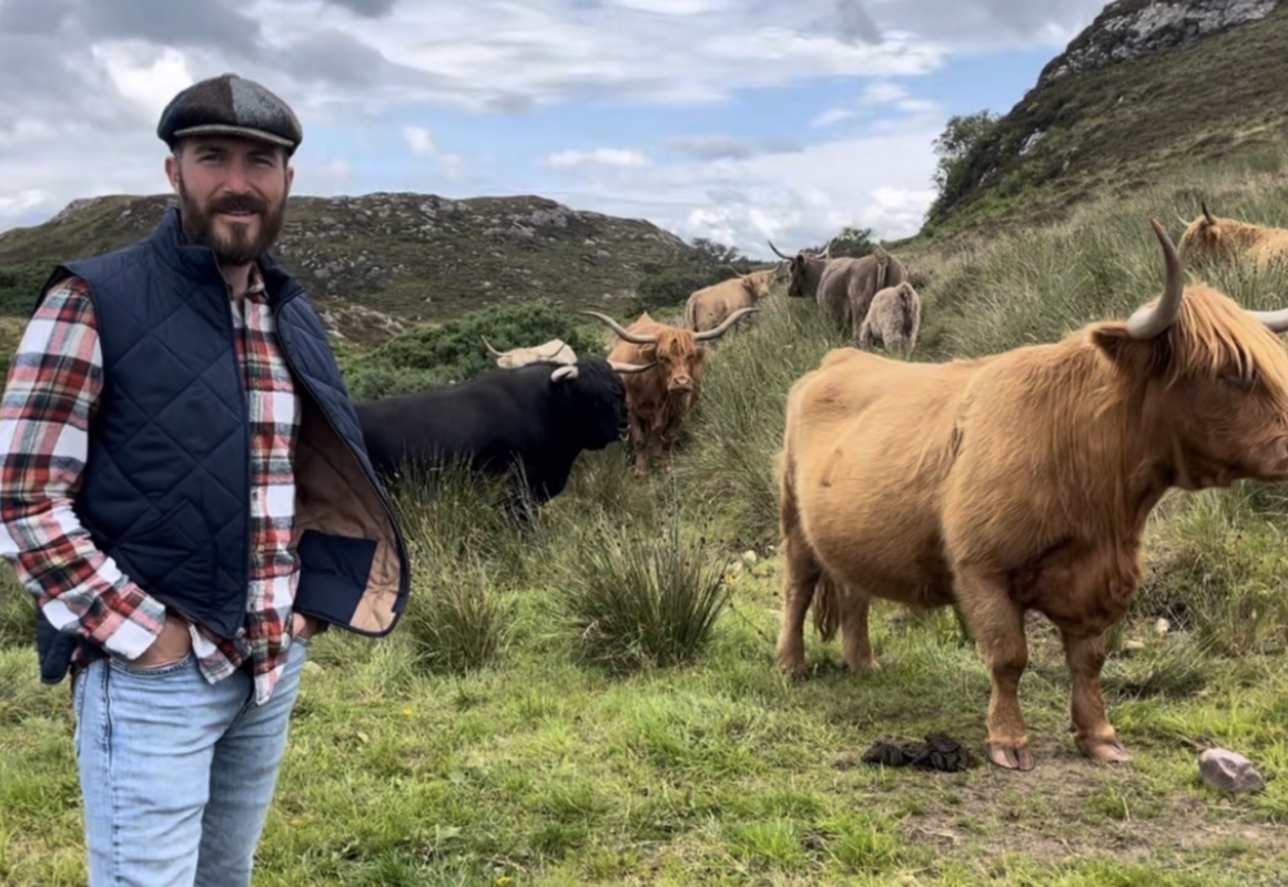 Man wearing a flat cap, plaid shirt, and navy vest standing beside a herd of Highland cows grazing on a grassy hillside in a rugged, rural landscape.