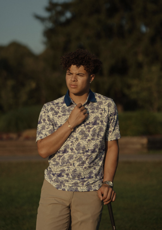 Golfer wearing a patterned polo with a navy collar and khaki pants standing outdoors at golden hour, adjusting his collar while holding a golf club on a lush course.