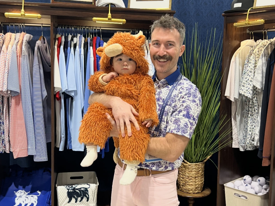 Man standing in the Zipelly booth at the PGA Show 2026 holding a baby dressed in a fuzzy orange Highland cow costume, surrounded by neatly displayed shirts, wooden fixtures, and branded merchandise.