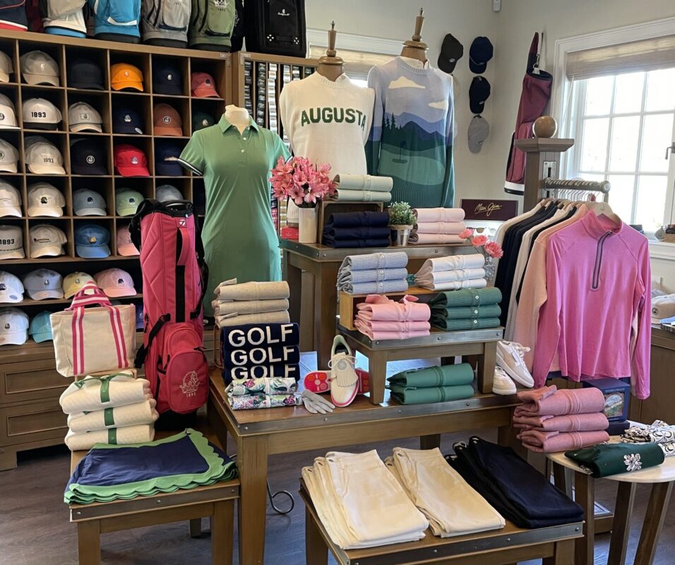 Golf shop display featuring folded apparel, mannequins, and accessories in green and pink tones with Augusta-themed sweaters and signage