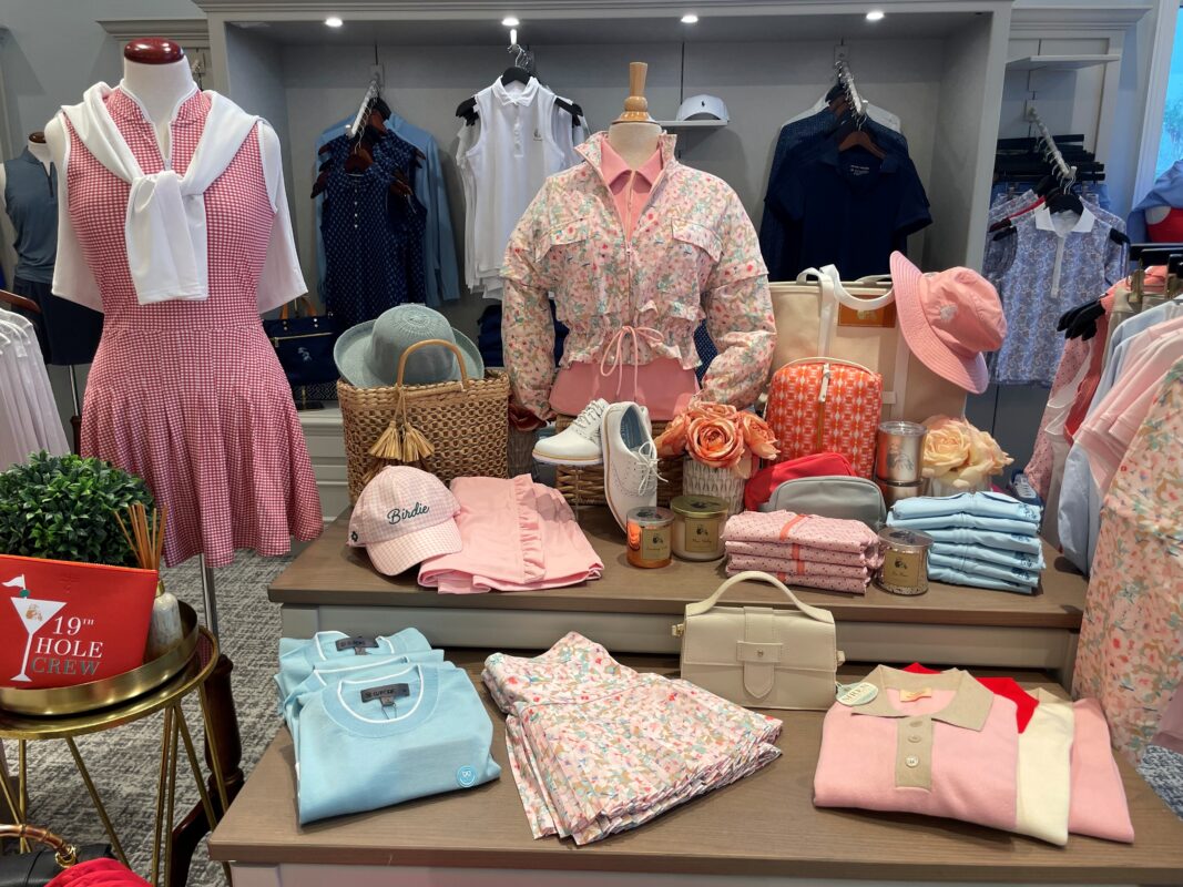 Merchandised golf shop display featuring coordinated pastel apparel, floral prints, accessories, and handbags arranged on table and mannequins