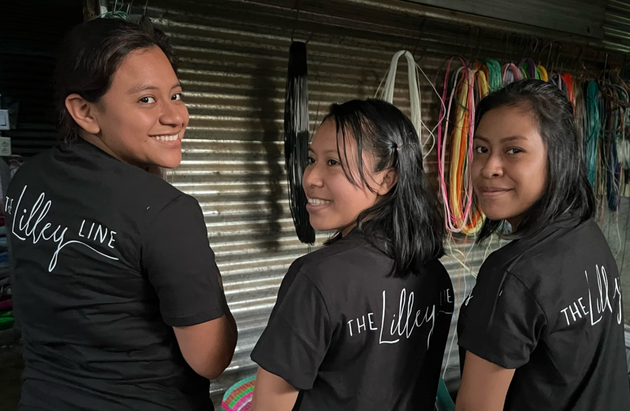 Three female artisans smiling and standing together in a workshop, wearing matching shirts with “The Lilley Line” logo and colorful weaving cords in the background