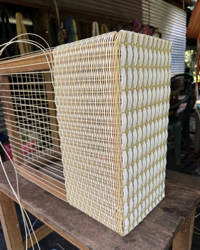 Close-up of a woven tote bag in progress on a loom, showing cream and white fibers being handwoven in a workshop setting