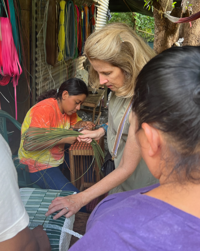 Woman observing and learning from artisans weaving by hand on a loom, with colorful cords and materials hanging in a workshop setting