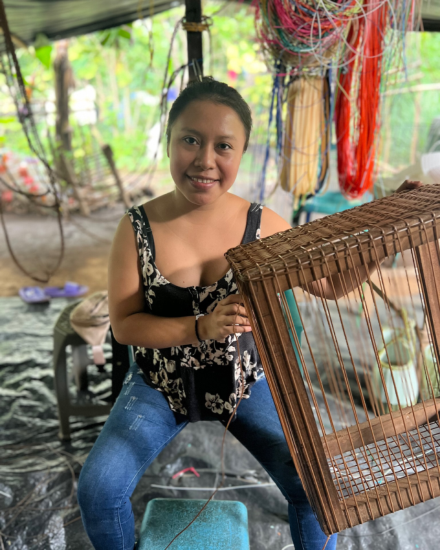 Woman seated and smiling while weaving a wicker basket by hand, with colorful cords and tools visible in the background.