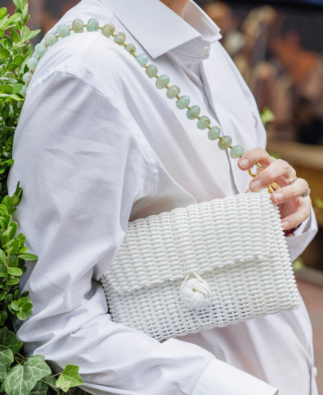 Close-up of a person holding a white woven clutch purse with a jade bead strap, wearing a white shirt with greenery in the background