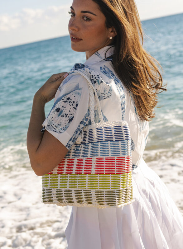 Woman standing on a beach wearing a white dress and holding a woven tote bag with blue, red, and green stripes, with ocean waves in the background