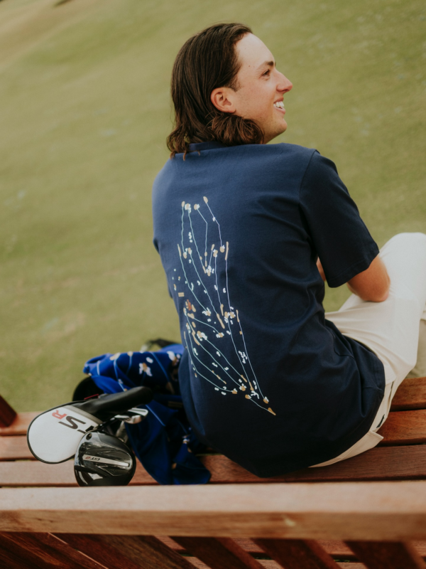 Man sitting on a wooden bench beside a golf course wearing a navy golf T-shirt with a floral graphic on the back, with golf clubs resting nearby.