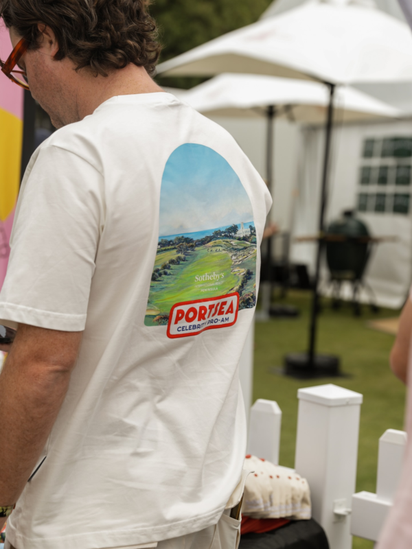 Man wearing a white T-shirt with a large golf course graphic and “Portsea Celebrity Pro-Am” text on the back, standing outdoors near umbrellas and event tents.