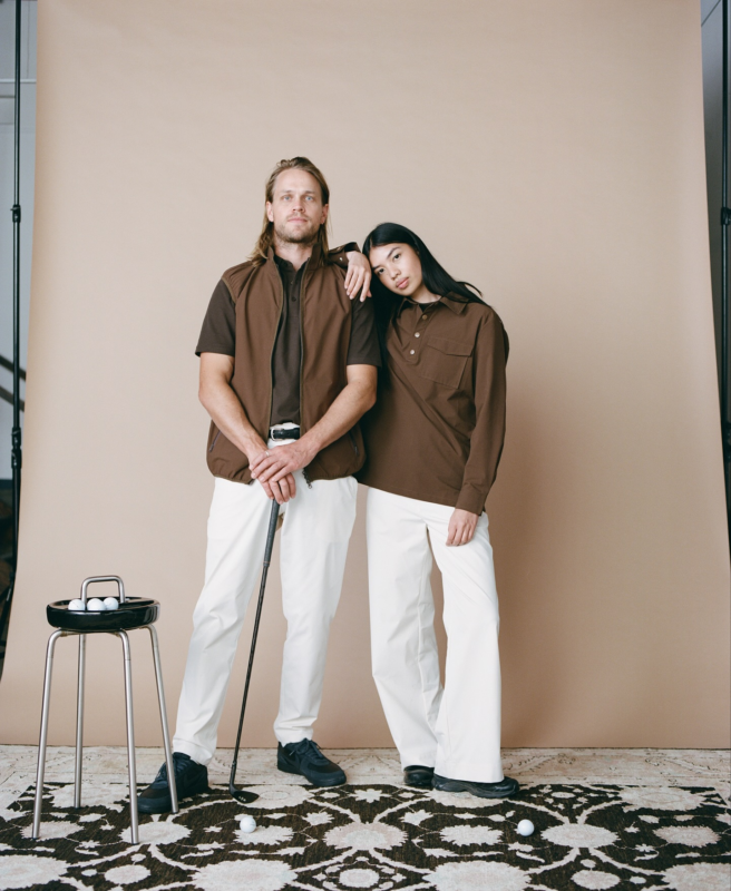 Male and female models wearing brown golf apparel and white pants stand together in a studio setting with a golf club, stool holding golf balls, and neutral backdrop.