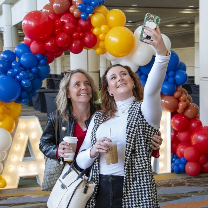 Two women smile for a selfie in front of a vibrant red, yellow, and blue balloon arch. Both hold coffee cups, with the woman on the right wearing a checkered vest over a white long-sleeve shirt.