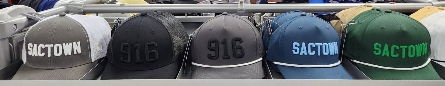 Row of “SacTown” and “916” embroidered baseball caps in various colors displayed on a retail shelf at Haggin Oaks Golf Super Shop.
