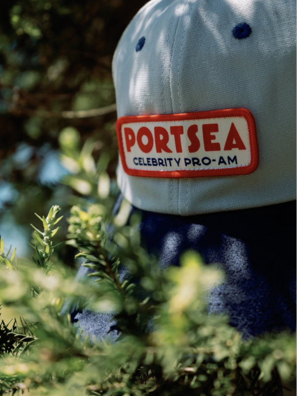 Close-up of a light-colored golf cap with a red patch reading “Portsea Celebrity Pro-Am,” photographed among green foliage outdoors.