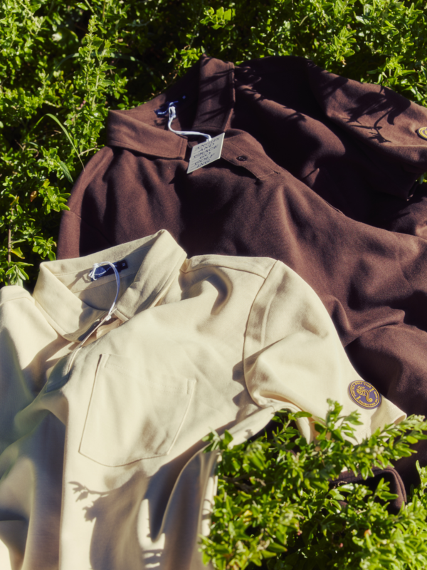 Two golf outer layers, one brown and one cream, laid on green foliage outdoors with visible collar details and embroidered sleeve patches.