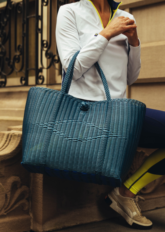 Person wearing a white athletic top and navy leggings holding a large blue woven tote bag while seated on stone steps