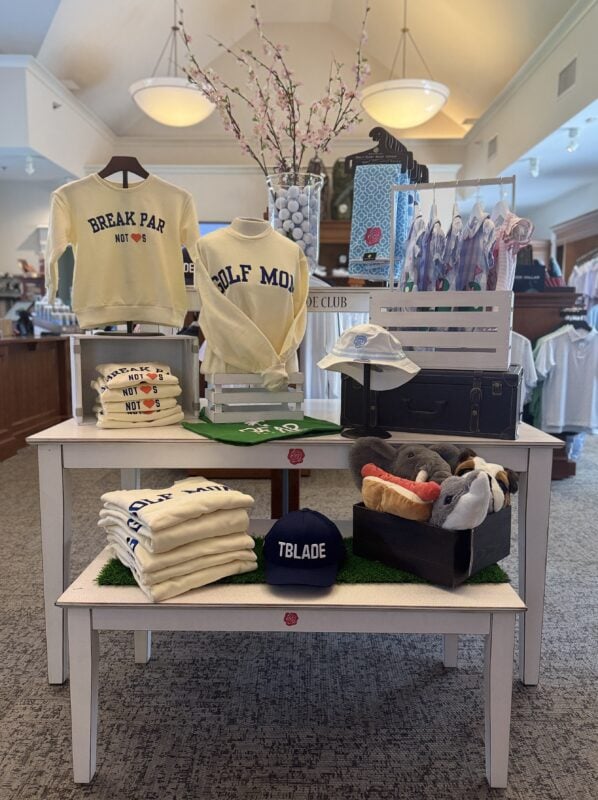 Mother’s Day golf shop display featuring cream sweatshirts with “Break Par Not Hearts” and “Golf Mom” text, folded apparel, children's toys and apparel, hats, and women's accessories arranged on tiered white tables with pink cherry blossom branches in a vase, creating a soft, elevated spring presentation.