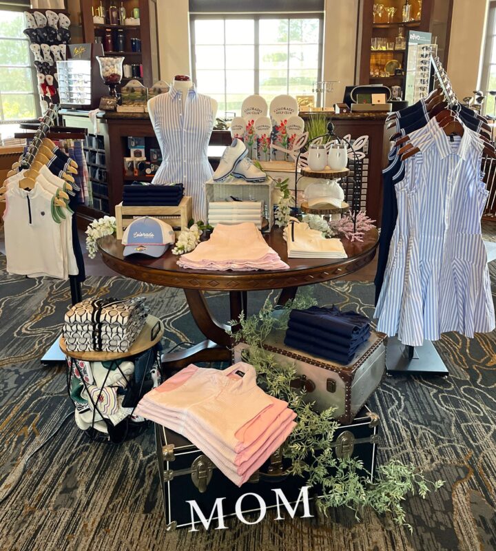 Round table Mother’s Day display with coordinated women’s golf apparel in soft pink, blue, and navy tones, styled with hats, shoes, and accessories; a vintage trunk labeled “MOM” anchors the display alongside florals and layered product groupings.