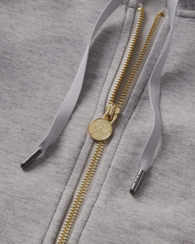 Close-up of a grey hoodie showing a gold zipper with a branded pull and matching drawstrings.