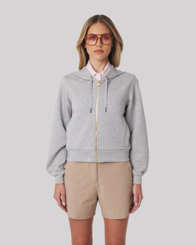 Female model facing forward in a grey zip-up hoodie layered over a collared shirt, paired with tan shorts.