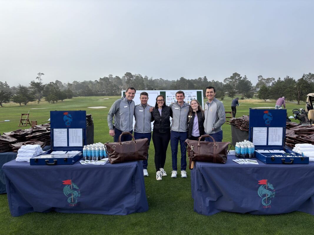 Group of six staff members standing on a golf course at Monterey Peninsula Country Club, smiling between two display tables set with branded merchandise, leather duffel bags, and bottled items, with the driving range in the background