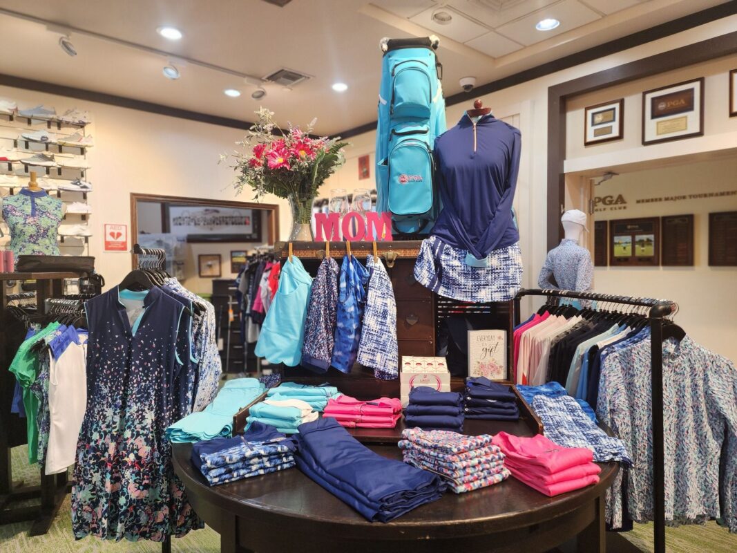 Golf shop merchandising display centered around a “MOM” sign, featuring coordinated women’s apparel in blue, teal, and pink tones, folded and hung in layered arrangements with a golf bag, florals, and accessories creating height and visual balance.