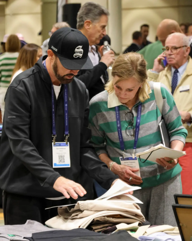 Two retail professionals wearing event badges review folded apparel on a display table at a busy trade show, discussing product details while referencing a notebook.