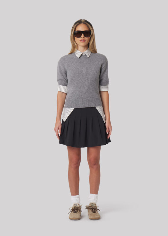 Female model standing front-facing in a grey short-sleeve sweater layered over a collared shirt, paired with a black pleated skirt and sneakers.