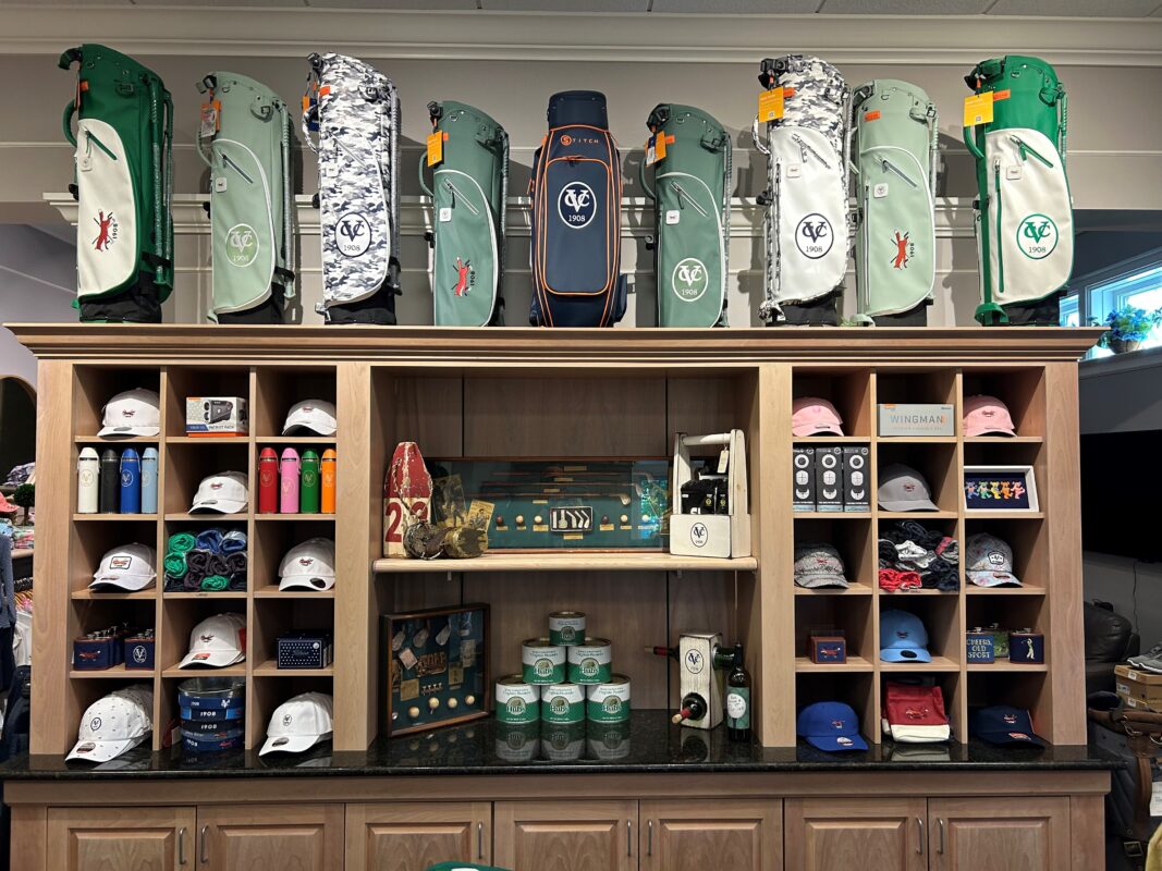Retail display inside a golf shop featuring neatly arranged hats, accessories, and drinkware on wooden shelving, with multiple golf bags displayed across the top shelf and curated merchandise organized by color and category
