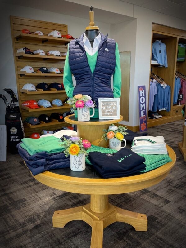 Round table Mother’s Day display featuring a mannequin styled in a green top and navy vest, surrounded by folded apparel and accessories, with floral arrangements in mugs labeled “Mom,” “Dog Mom,” and “Grandma,” and a small sign highlighting Mother’s Day, creating a personalized and gift-focused presentation.