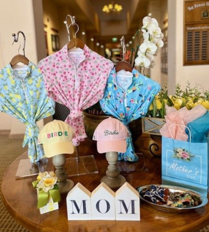 Boutique-style Mother’s Day display with floral-print women’s golf tops, pastel hats, a gift bag labeled “Mother’s Day,” and decorative flowers arranged on a round table with small “MOM” blocks, creating a gift-ready, intimate presentation.